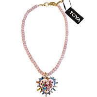 A Tova brand beaded necklace featuring light pink faceted beads, a gold-tone clasp, and a large, colorful heart-shaped pendant with cartoon-style illustrations and rhinestones.