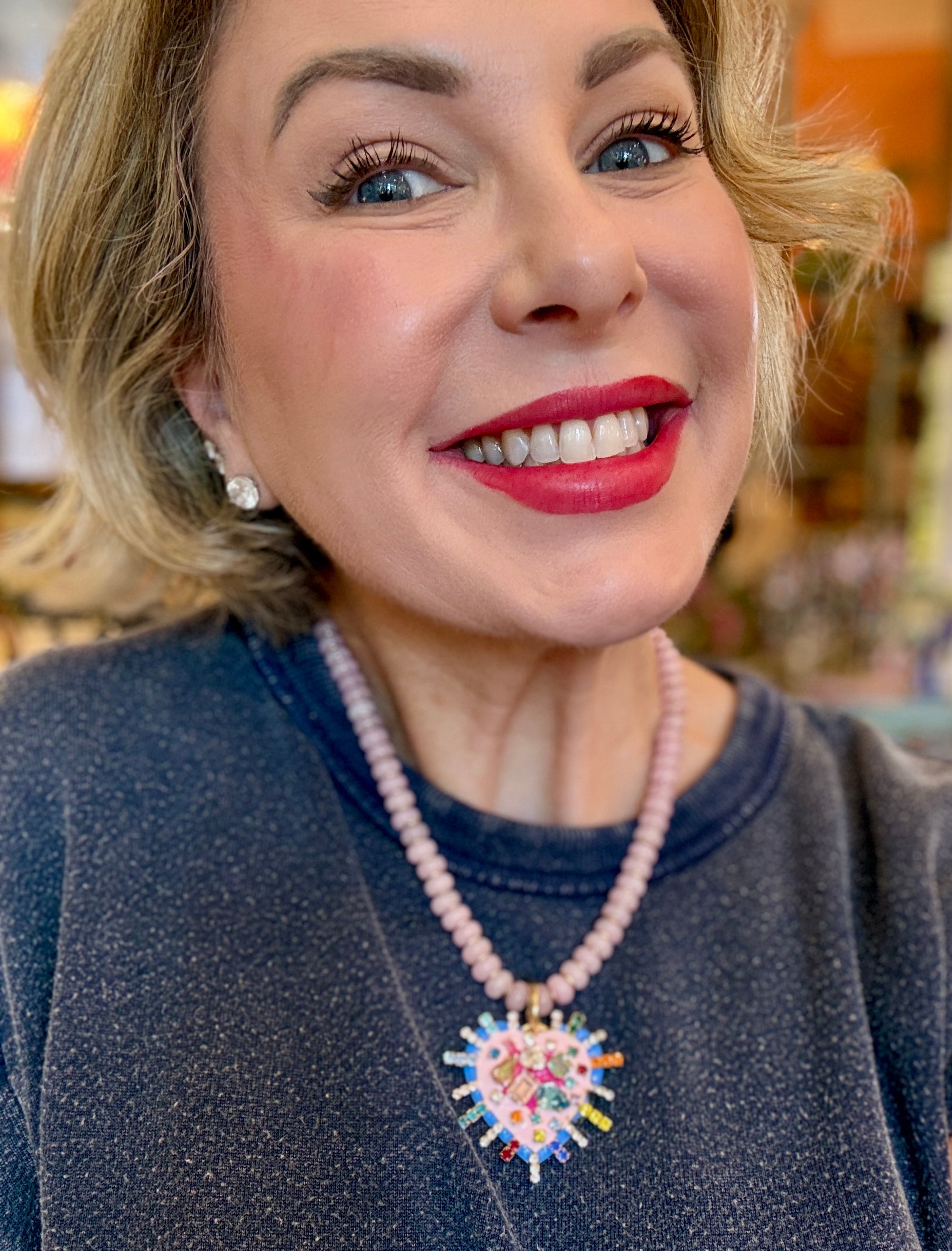 A Tova brand beaded necklace featuring light pink faceted beads, a gold-tone clasp, and a large, colorful heart-shaped pendant with cartoon-style illustrations and rhinestones.