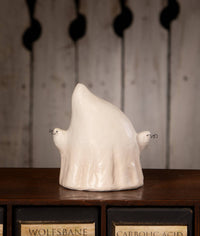 A ceramic ghost figurine in a glossy off-white glaze, displayed on a dark wooden surface with vintage apothecary-style drawers in the background.