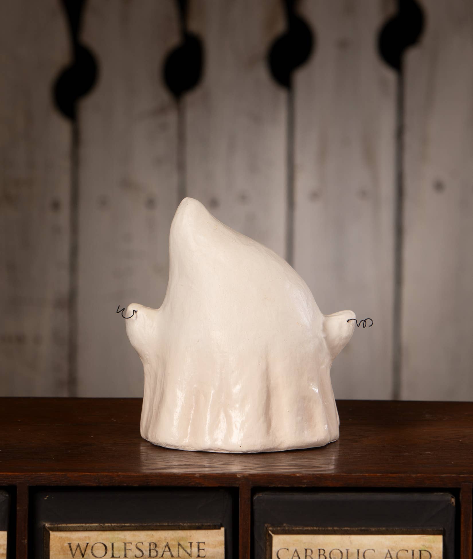 A ceramic ghost figurine in a glossy off-white glaze, displayed on a dark wooden surface with vintage apothecary-style drawers in the background.