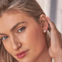 Close-up of a woman wearing a small round stud earring with a clear gemstone, with her hand near her ear against a soft light gray background.