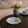 Light blue ceramic dish with decorative elements on a dark wooden table, with a vase of white flowers in the background.