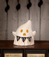 A white ceramic ghost-shaped cookie jar with yellow eyes and mouth, holding a black bunting banner that says 'BOO', displayed on a dark wooden shelf.