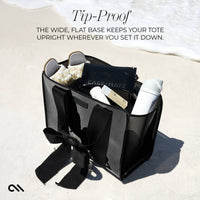 Black tote bag with items on a sandy beach, featuring text about its tip-proof design.