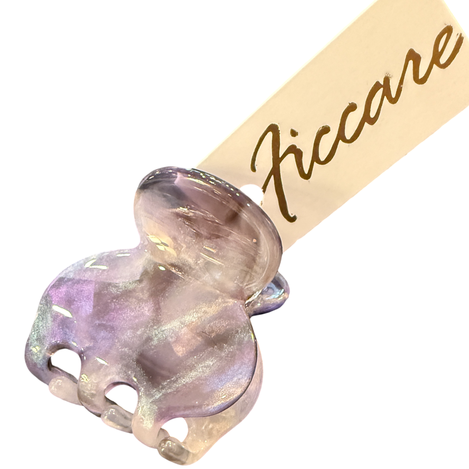 A close-up of a decorative hair claw clip with an iridescent lavender and pink elephant head charm, attached to a beige paper tag printed with the brand name 'Ficcare' in gold script, set against a plain white background.