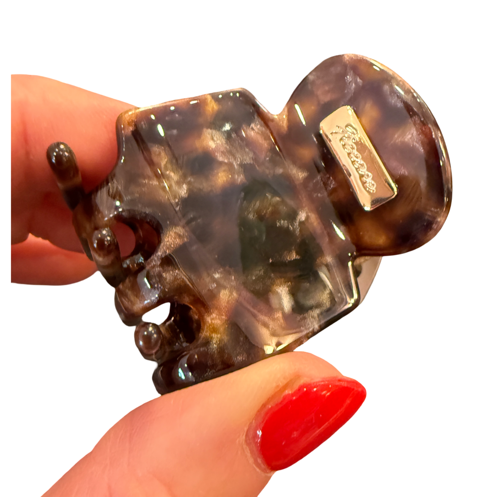 A tortoiseshell hair claw clip held by a hand with red nail polish against a plain white background. The brand name 'Sartoso' is visible on a small metal tag attached to the clip.