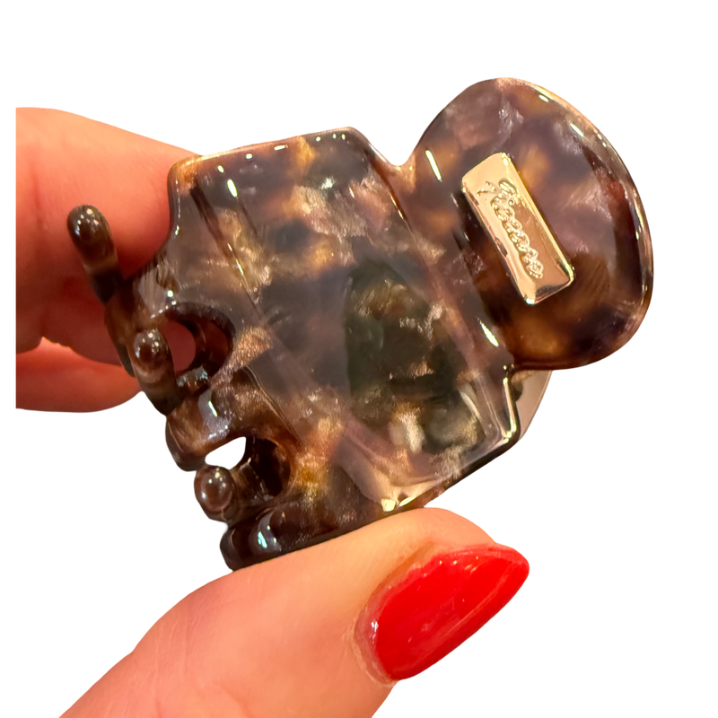 A tortoiseshell hair claw clip held by a hand with red nail polish against a plain white background. The brand name 'Sartoso' is visible on a small metal tag attached to the clip.