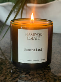 A lit amber glass jar candle labeled 'Flaming Estate Banana Leaf' with a visible flame, photographed on a speckled countertop.