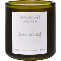 An 8.0 oz glass candle in a dark olive green jar with a white paper label reading 'FLAMING ESTATIE Banana Leaf'.
