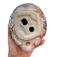 The underside of a vintage ceramic clown figurine held in a hand, showing the glazed pink and white face, decorative floral side details, and a white manufacturer label.