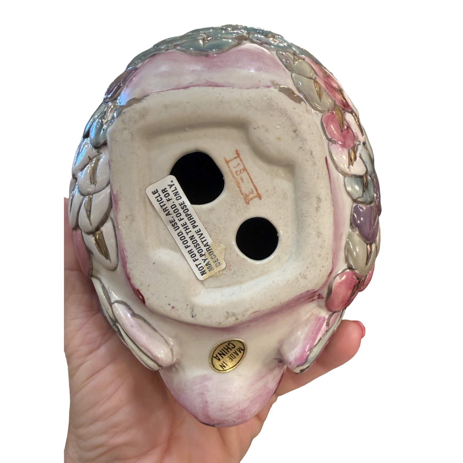 The underside of a vintage ceramic clown figurine held in a hand, showing the glazed pink and white face, decorative floral side details, and a white manufacturer label.