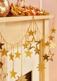 Gold metallic star garland hanging from chains across a light wood mantel, with additional holiday decorations visible above.