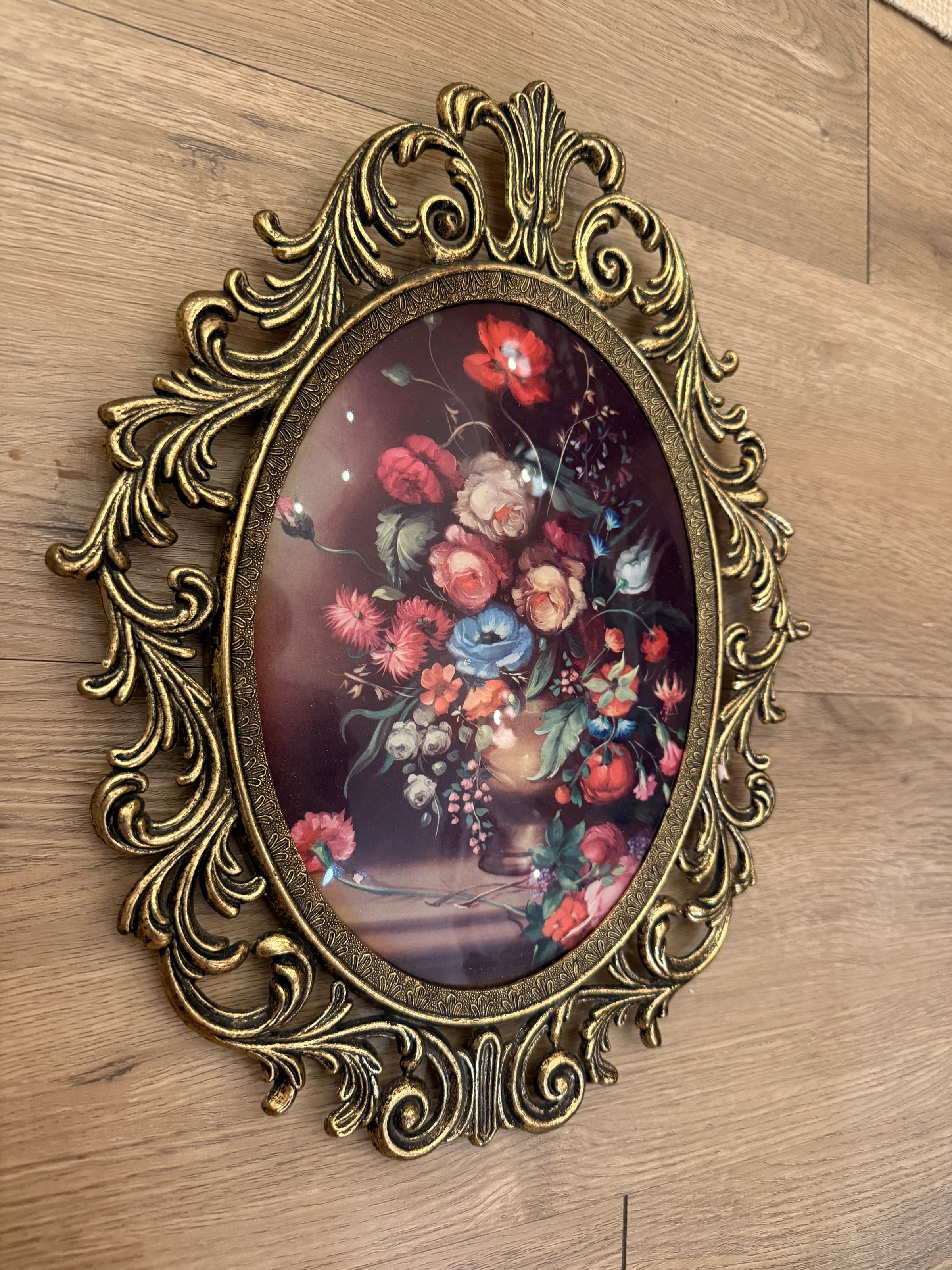 An ornate oval wall art piece with a distressed gold frame, featuring a colorful floral painting of red, pink, and blue flowers on a dark background, displayed on a light wood floor.