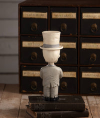 Vintage-style figurine wearing a top hat on stacked books with a wooden cabinet background.