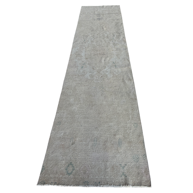 A long rectangular area rug in a distressed light gray color with subtle faded blue-gray and beige undertones, featuring a distressed traditional pattern and short fringe on the ends.
