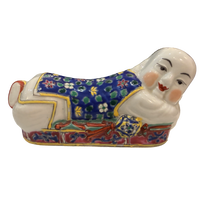 A ceramic figurine of a reclining figure in traditional East Asian-style clothing, decorated with a blue floral pattern, red and gold details, and a white face and hands, isolated on a white background.