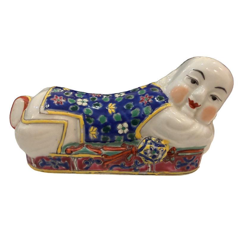 A ceramic figurine of a reclining figure in traditional East Asian-style clothing, decorated with a blue floral pattern, red and gold details, and a white face and hands, isolated on a white background.