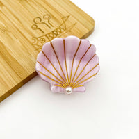 Pink shell-shaped object with gold accents and a pearl on a wooden surface