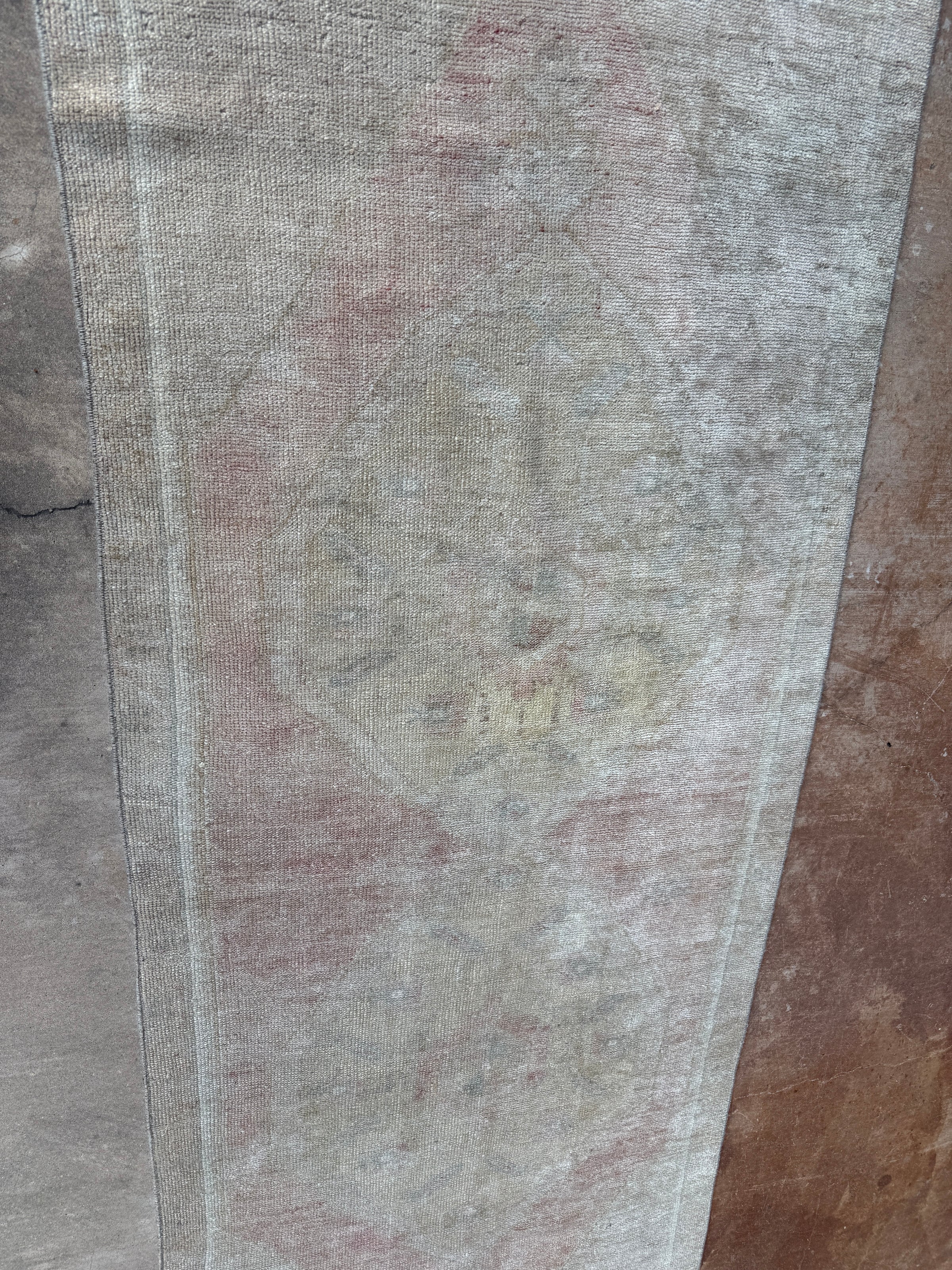 A close-up of a distressed area rug with a faded traditional pattern in muted tones of gray, beige, and dusty pink, laid flat on a concrete floor.