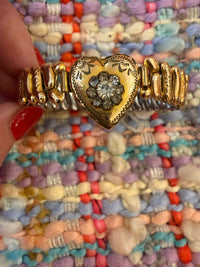 A gold-toned vintage-style bangle bracelet with a heart-shaped center and a floral motif, held by a hand with red nail polish against a colorful textured knit background.