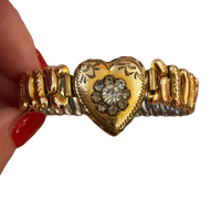 A gold heart-shaped cuff bracelet with a central diamond cluster, held by a hand with red nail polish against a white background.