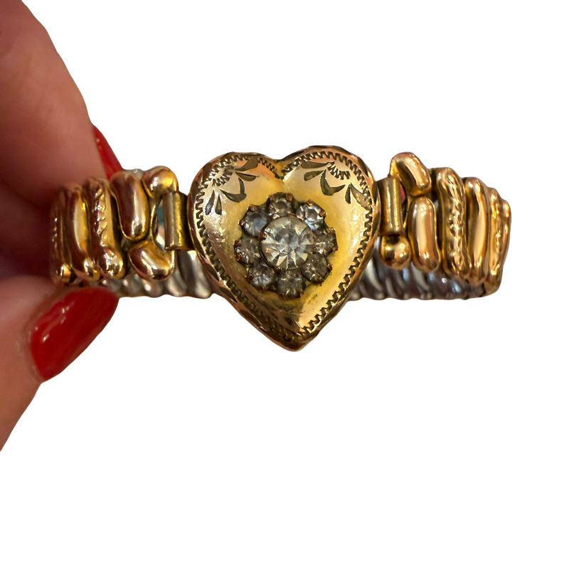 A gold heart-shaped cuff bracelet with a central diamond cluster, held by a hand with red nail polish against a white background.