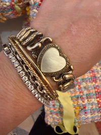 A close-up of a person's wrist wearing a stack of chunky vintage-style bracelets, including a heart-shaped charm bracelet, a rope chain bracelet, and a silver-toned chain bracelet with clear stones.