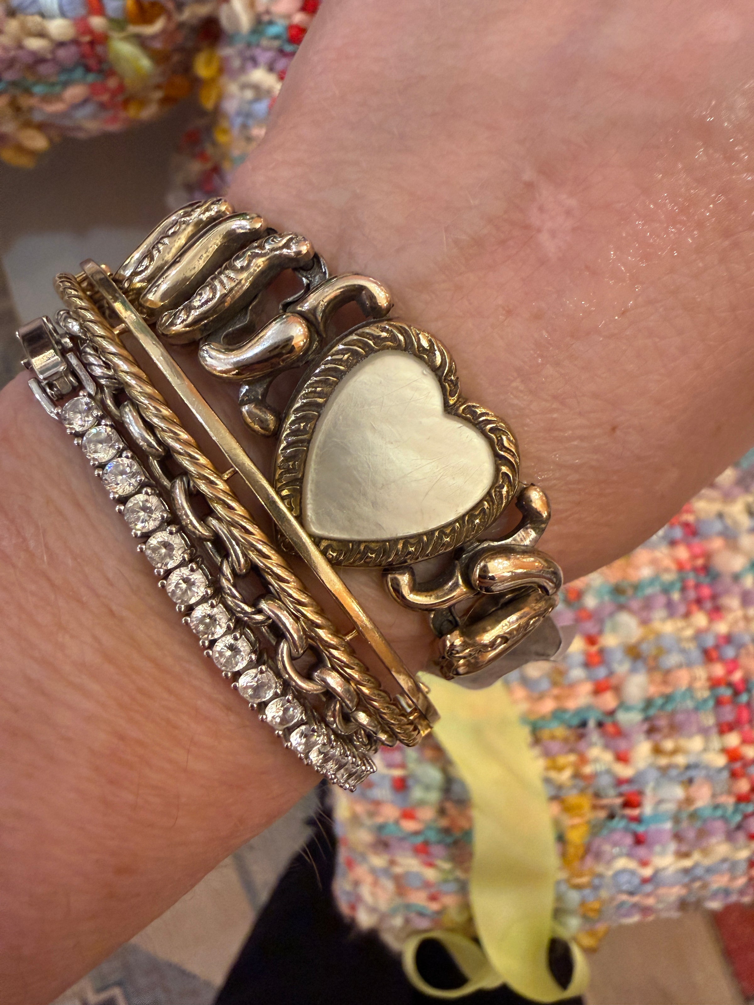 A close-up of a person's wrist wearing a stack of chunky vintage-style bracelets, including a heart-shaped charm bracelet, a rope chain bracelet, and a silver-toned chain bracelet with clear stones.