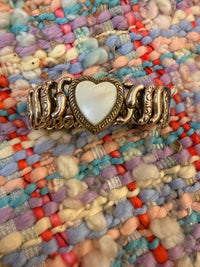 A wide gold-toned metal cuff bracelet featuring a central heart-shaped inlay with a white mother-of-pearl finish, displayed on a colorful, textured boucle fabric background.
