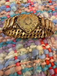 A gold-toned metal bracelet featuring a central heart-shaped charm with an engraved heart and floral details, flanked by textured twisted links, displayed on a colorful woven rug.