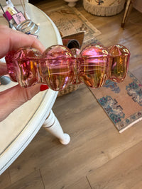 A hand holding a chunky, glossy pink glass hair claw clip in a home setting with a wooden floor and a white table visible.