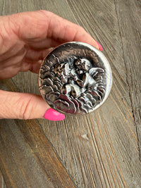 Silver round object with engraved design held by a hand on a wooden surface