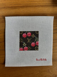 Cross-stitched design on fabric with 'Bad Bitch' text, placed on a wooden surface.