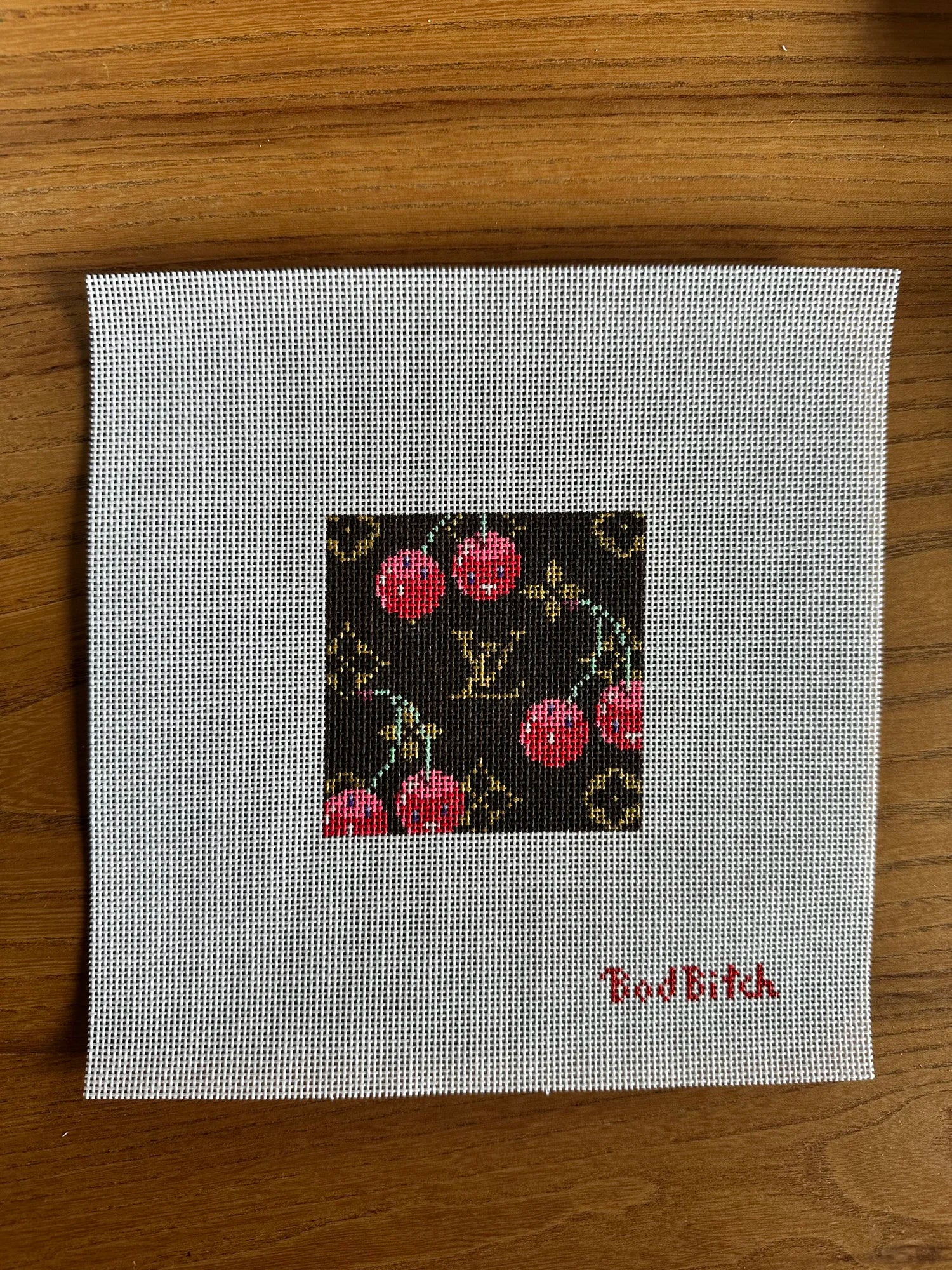 Cross-stitched design on fabric with 'Bad Bitch' text, placed on a wooden surface.