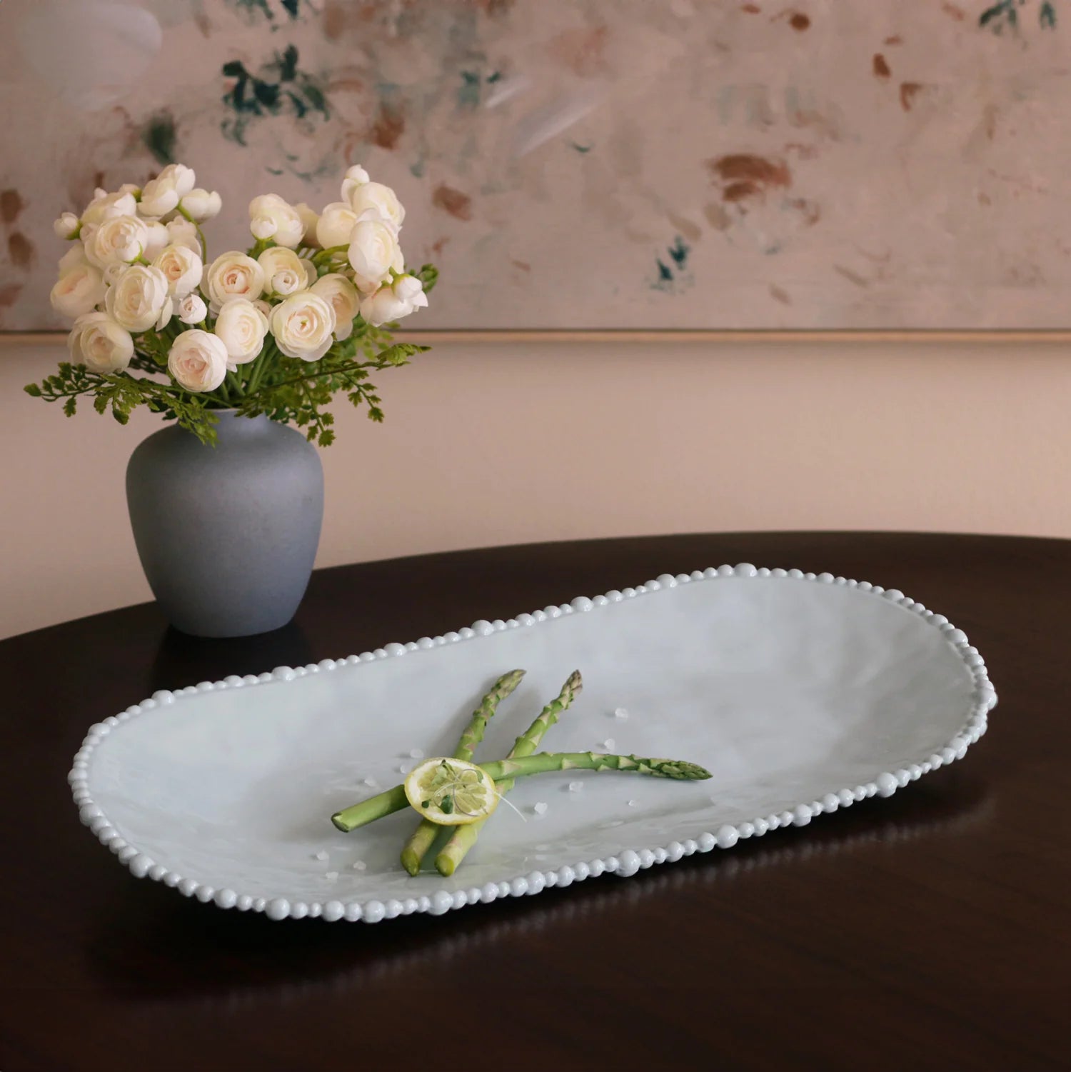 Decorative tray with asparagus and lemon on a table with a vase of flowers in the background.
