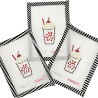 Four cross-stitched designs of a milkshake with red straw on white fabric, framed by black and white striped borders.