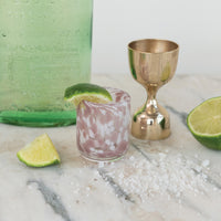 Cocktail setup with a pink glass, gold shaker, and lime wedges on a marble surface.