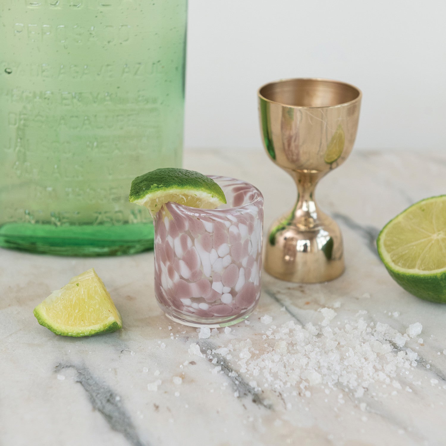 Cocktail setup with a pink glass, gold shaker, and lime wedges on a marble surface.