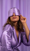 Woman wearing a purple satin robe and matching sleep mask against a purple curtain.
