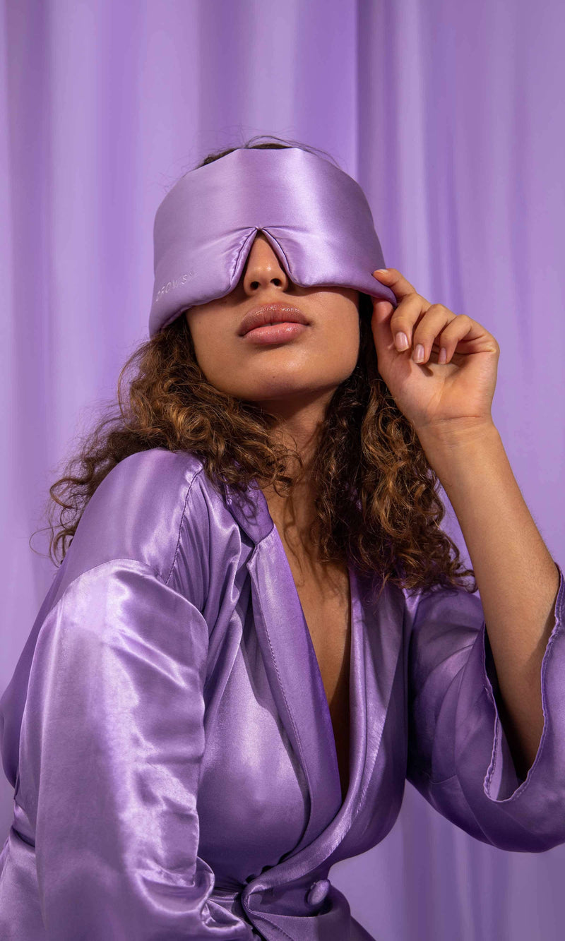Woman wearing a purple satin robe and matching sleep mask against a purple curtain.