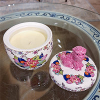 Decorative container with a pink dog figurine on a floral lid, placed on a glass surface.