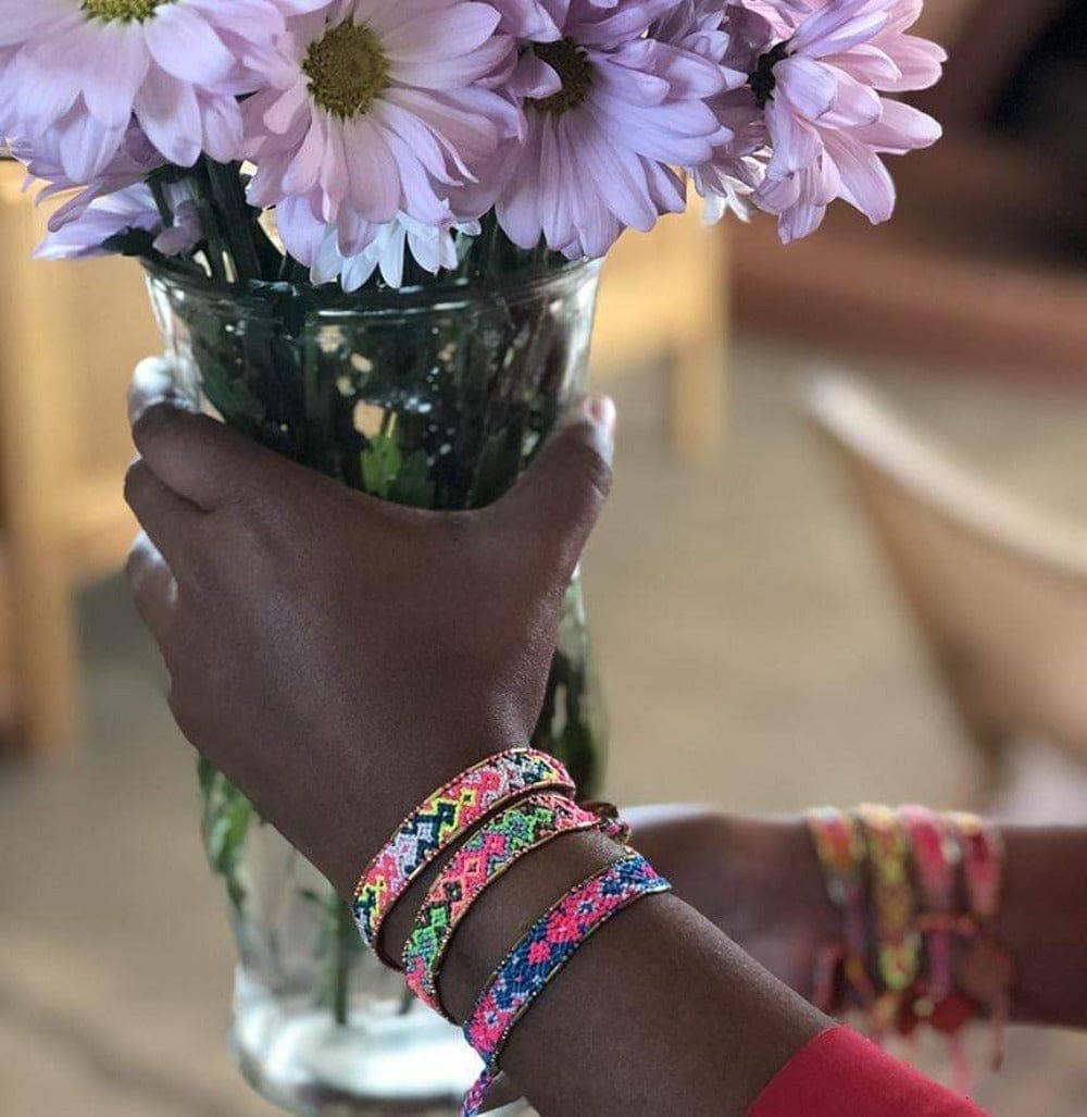Hand holding a vase of pink flowers with colorful bracelets on a blurred background