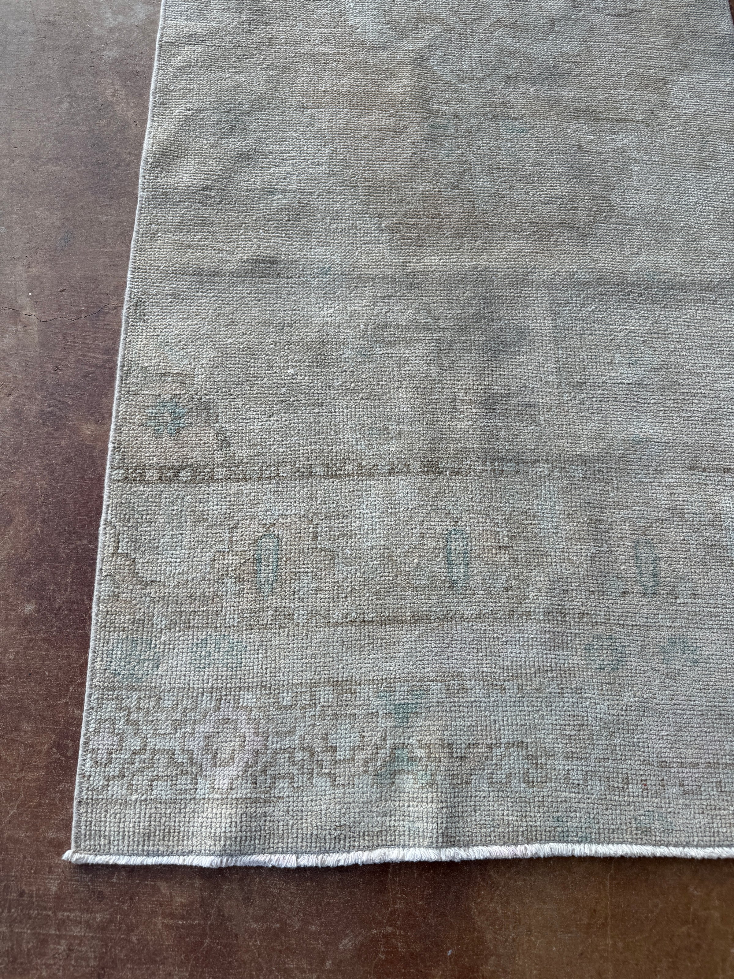 A close-up of a distressed area rug with a faded taupe-gray and muted blue-green pattern, laid flat on a dark brown wooden floor.