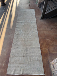 A long, narrow runner rug laid out on a concrete floor, photographed from above at an angle. The rug has a distressed, vintage appearance with faded beige, gray, and muted blue-gray tones and subtle traditional patterns near the bottom.