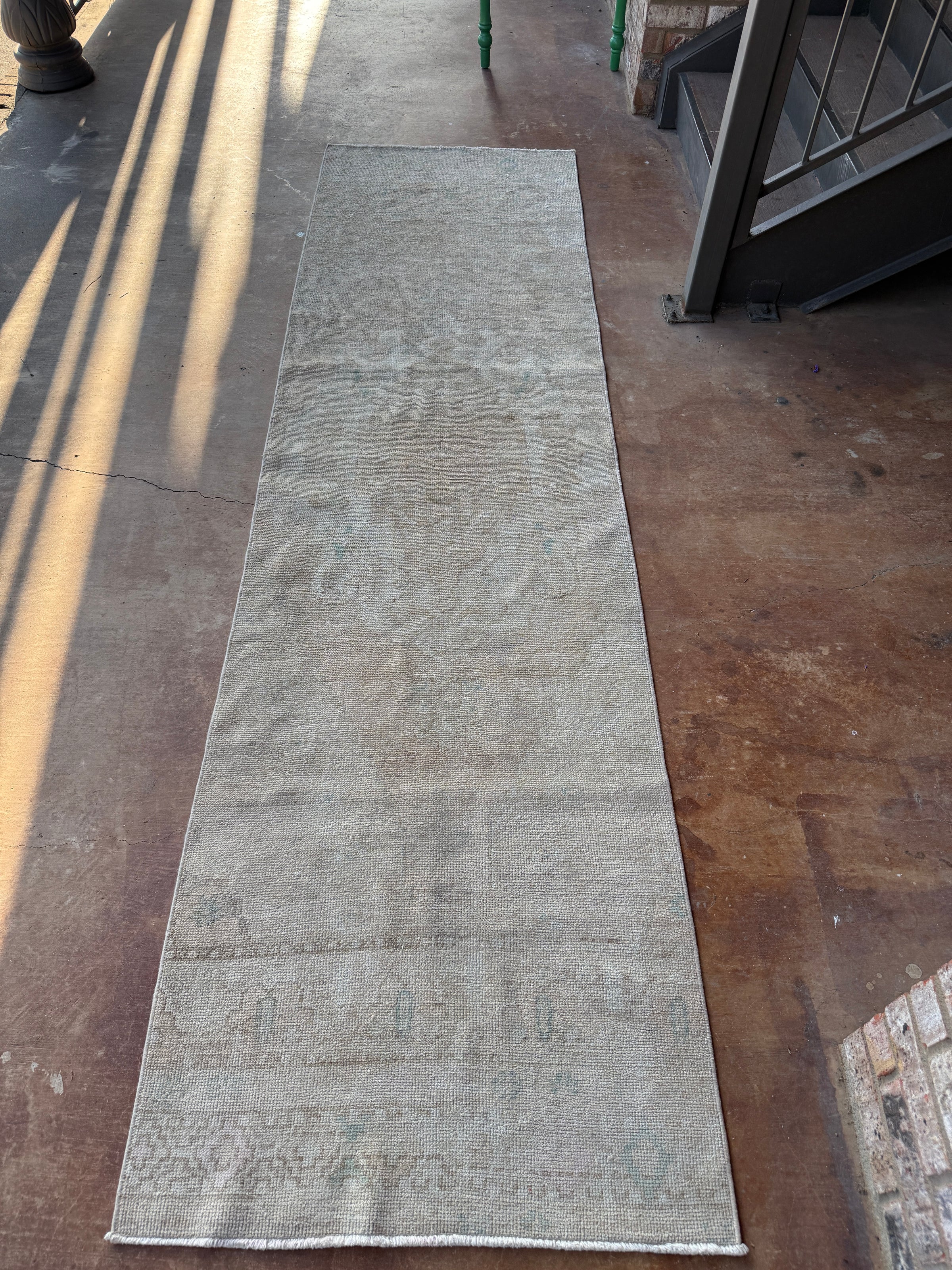 A long, narrow runner rug laid out on a concrete floor, photographed from above at an angle. The rug has a distressed, vintage appearance with faded beige, gray, and muted blue-gray tones and subtle traditional patterns near the bottom.