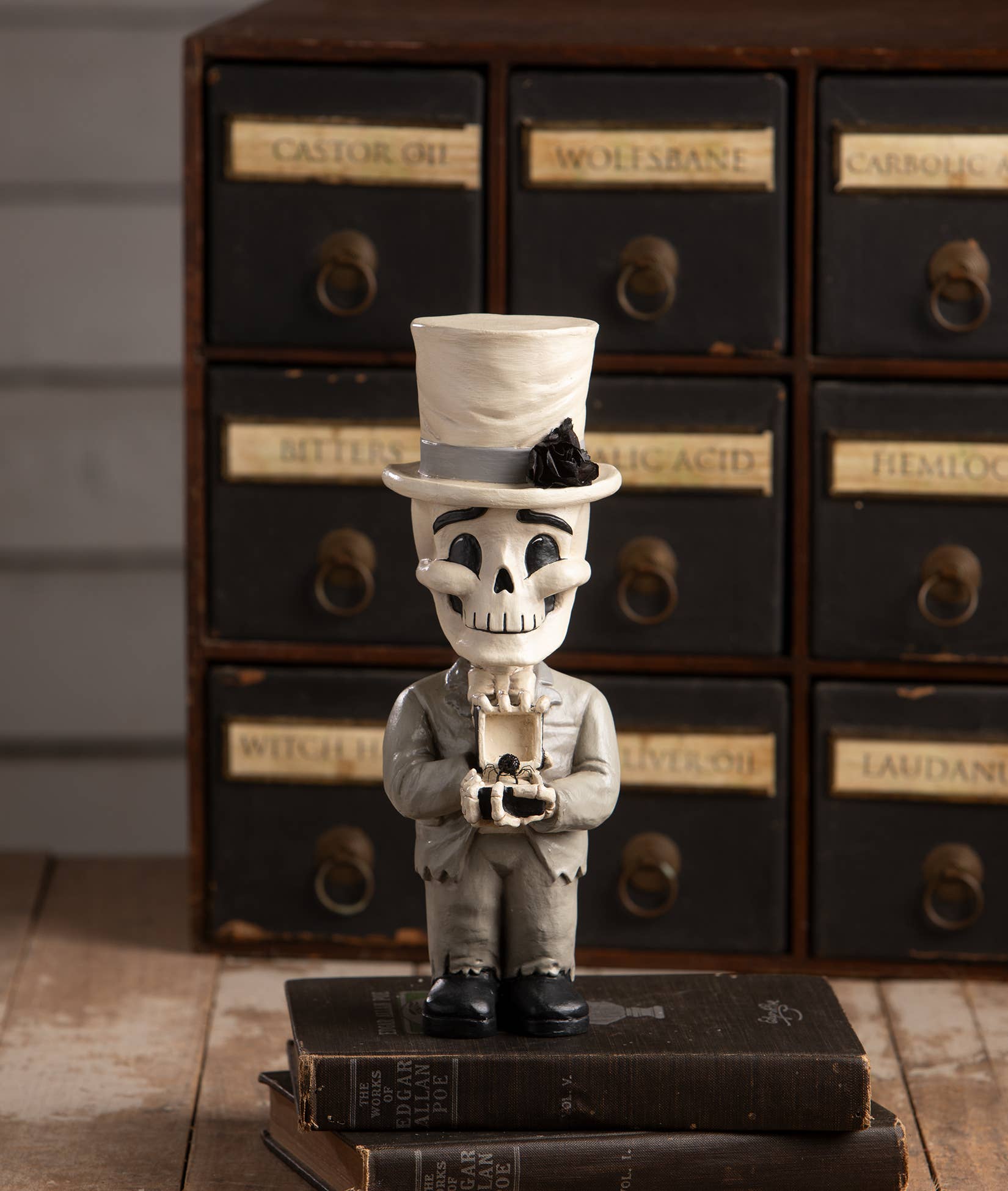 Skull figurine in a top hat and suit standing on books with vintage apothecary drawers in the background.