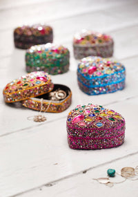 A collection of small, heart-shaped jewelry boxes covered in colorful rhinestones and sequins, arranged on a white wooden surface. One box is open, revealing rings inside, while others are closed.