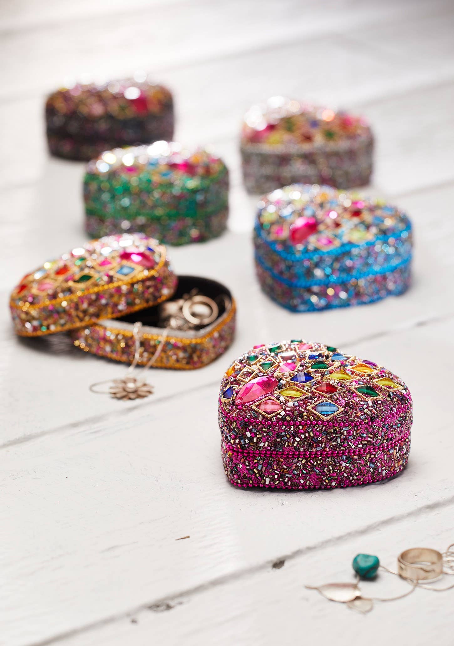 A collection of small, heart-shaped jewelry boxes covered in colorful rhinestones and sequins, arranged on a white wooden surface. One box is open, revealing rings inside, while others are closed.