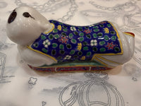 A ceramic figurine of a dog wearing a colorful patterned robe, resting on a white fabric with gray line drawings.