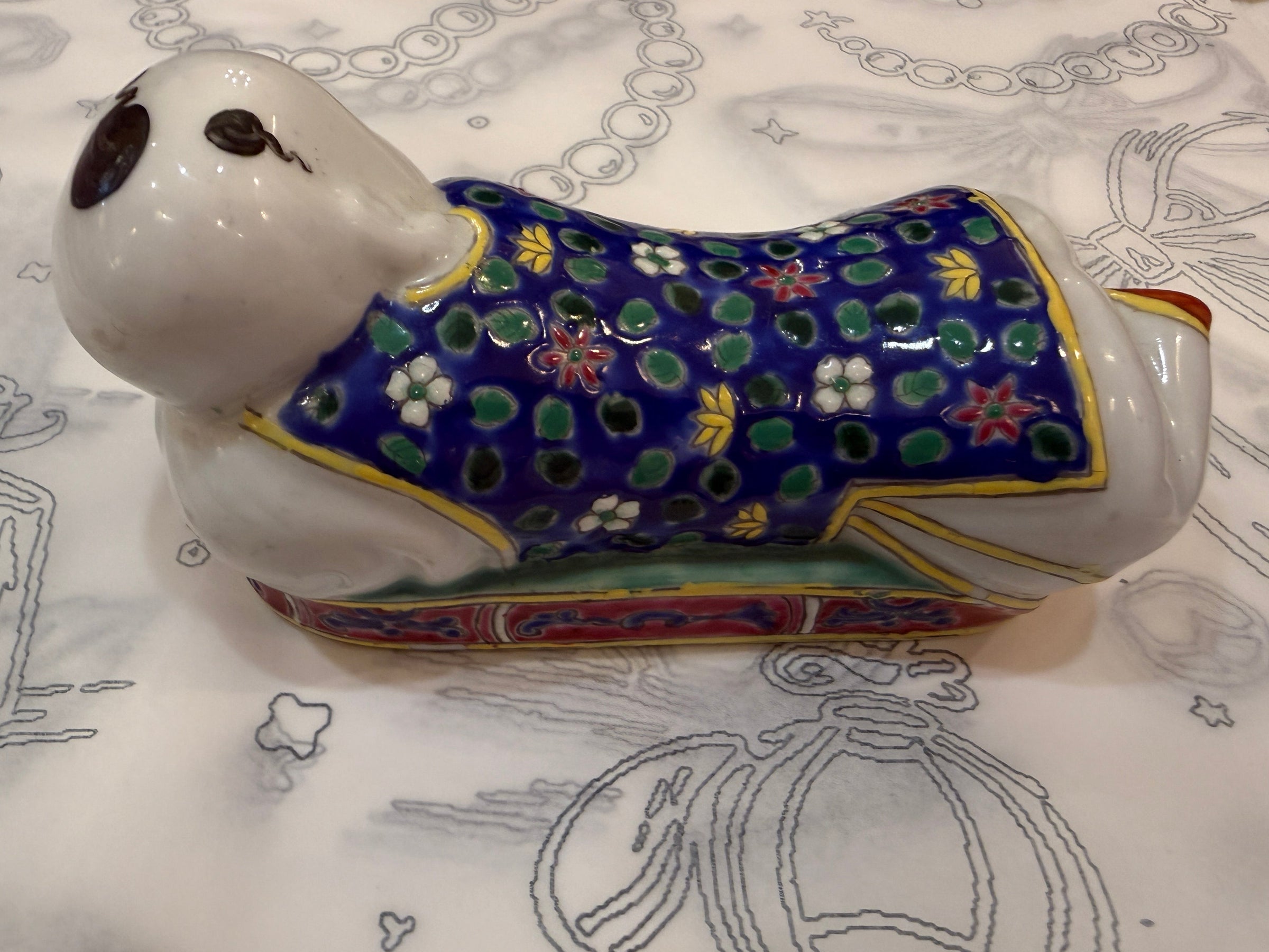 A ceramic figurine of a dog wearing a colorful patterned robe, resting on a white fabric with gray line drawings.